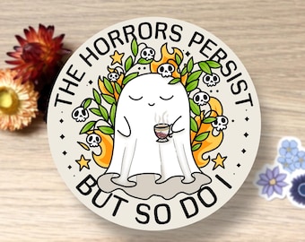 May include: A round sticker featuring a cartoon ghost holding a cup of coffee. The ghost is surrounded by flames, skulls, and leaves. The text "THE HORRORS PERSIST BUT SO DO I" is printed around the edge of the sticker.