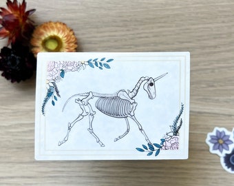 May include: A white card with a black and white illustration of a unicorn skeleton surrounded by blue and pink flowers.