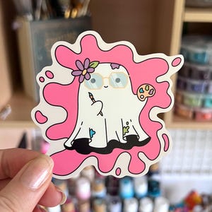 May include: A white ghost-shaped sticker with a pink paint splash design. The ghost wears glasses, a flower, and holds a paintbrush and palette. The sticker has a black outline and is held by a person.
