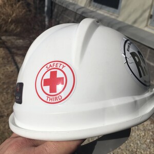 Puede incluir: Casco blanco con una cruz roja y las palabras "SAFETY THIRD" en una pegatina.