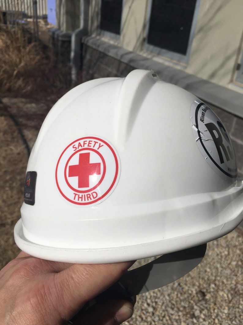 Puede incluir: Casco blanco con una cruz roja y las palabras "SAFETY THIRD" en una pegatina.
