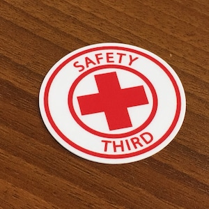 May include: A white round sticker with a red border. The sticker has a red cross in the center and the words "SAFETY THIRD" around the outside.