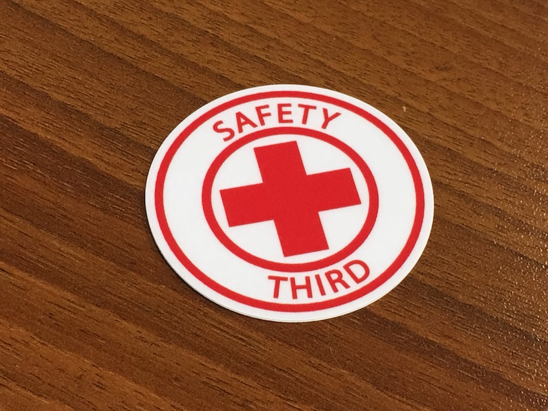 Puede incluir: Una pegatina redonda blanca con un borde rojo. La pegatina tiene una cruz roja en el centro y las palabras "SAFETY THIRD" alrededor.
