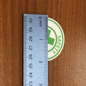 Puede incluir: Una pegatina blanca con una cruz verde y la palabra "SAFETY" en texto negro. La pegatina tiene un di&aacute;metro de aproximadamente 2,5 cm y se mide con una regla.