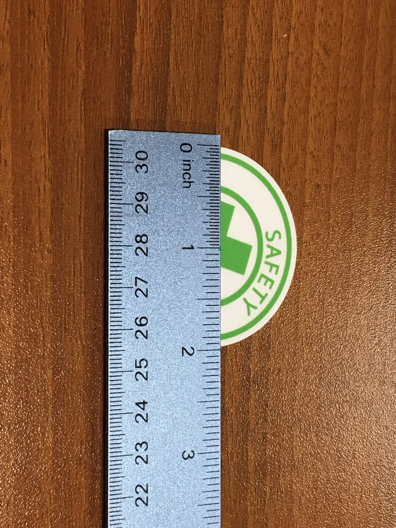 Puede incluir: Una pegatina blanca con una cruz verde y la palabra "SAFETY" en texto negro. La pegatina tiene un di&aacute;metro de aproximadamente 2,5 cm y se mide con una regla.