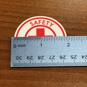 Puede incluir: Una pegatina redonda blanca con un borde rojo y la palabra "SAFETY" en letras rojas. Una cruz roja est&aacute; en el centro de la pegatina. La pegatina se mide con una regla, mostrando que tiene aproximadamente 2,5 cm de di&aacute;metro.