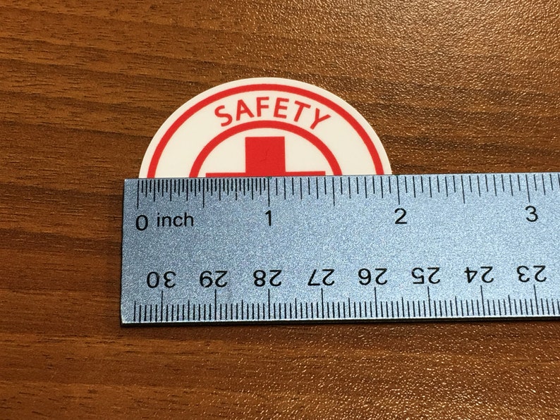 Puede incluir: Una pegatina redonda blanca con un borde rojo y la palabra "SAFETY" en letras rojas. Una cruz roja est&aacute; en el centro de la pegatina. La pegatina se mide con una regla, mostrando que tiene aproximadamente 2,5 cm de di&aacute;metro.