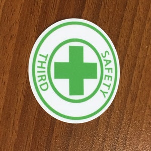 Puede incluir: Una pegatina redonda blanca con un borde verde. La pegatina tiene una cruz verde en el centro y las palabras "SAFETY THIRD" escritas alrededor del exterior.