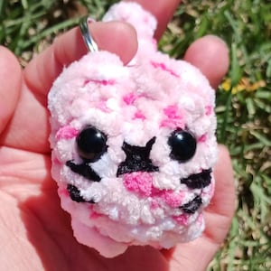 May include: A handmade pink and white plush keychain in the shape of a cartoon animal. The keychain has black eyes and black markings. The plush is held in a hand against a background of green grass.