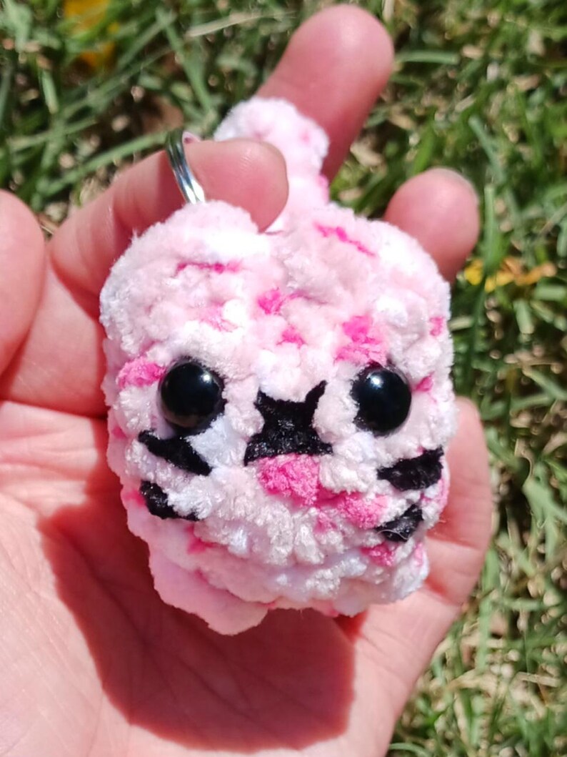 May include: A handmade pink and white plush keychain in the shape of a cartoon animal. The keychain has black eyes and black markings. The plush is held in a hand against a background of green grass.