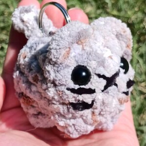 May include: A handmade, gray and brown crocheted cat keychain. The cat has black bead eyes and black stitched details for the nose and mouth. A silver metal keyring is attached to the top of the cat's head. The keychain is held in a person's hand.