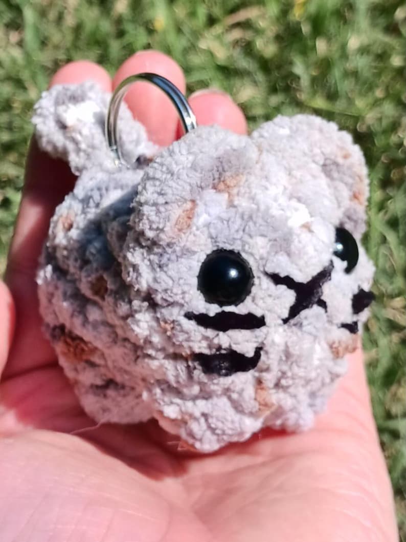 May include: A handmade, gray and brown crocheted cat keychain. The cat has black bead eyes and black stitched details for the nose and mouth. A silver metal keyring is attached to the top of the cat's head. The keychain is held in a person's hand.