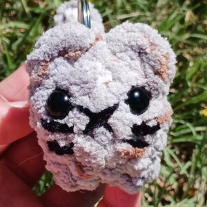 May include: A handmade, plush keychain in the shape of a cat's face. The cat has black eyes, a black nose, and black whiskers. The keychain is made of gray and brown yarn and has a silver key ring.
