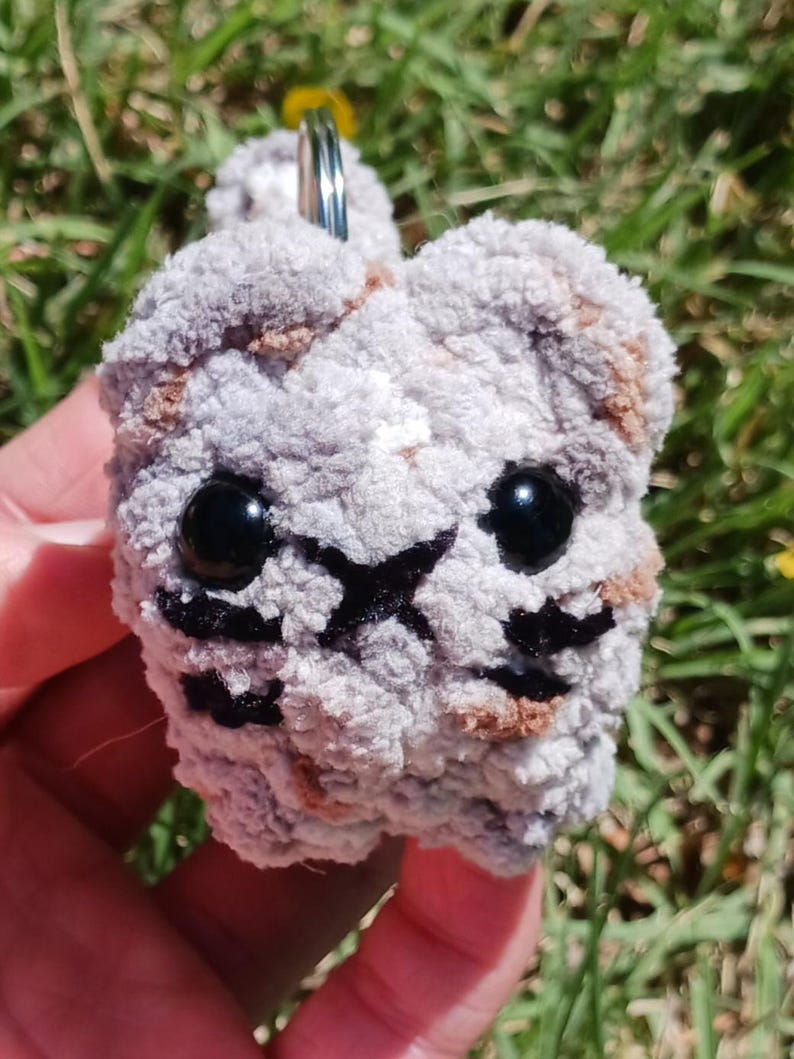 May include: A handmade, plush keychain in the shape of a cat's face. The cat has black eyes, a black nose, and black whiskers. The keychain is made of gray and brown yarn and has a silver key ring.
