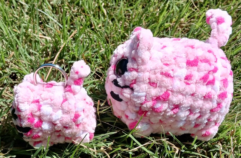 May include: Two handmade, pink and white crochet cat plushies with black facial details and large black eyes. One is a keychain, the other is a larger stuffed animal. Both are on green grass.