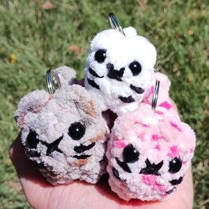 May include: Three handmade crochet keychains in the shape of small animals. One is white, one is pink, and one is gray. Each has black eyes and a black nose and mouth design. The keychains are held in a hand against a green background.