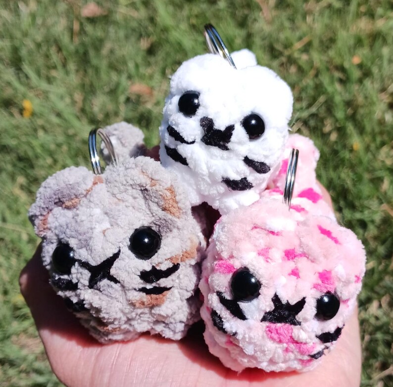 May include: Three handmade crochet keychains in the shape of small animals. One is white, one is pink, and one is gray. Each has black eyes and a black nose and mouth design. The keychains are held in a hand against a green background.