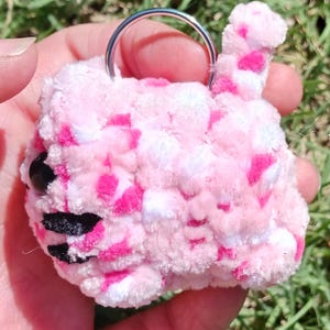 May include: A handmade, pink and white crochet keychain in the shape of a cat. The cat has black eyes and a black and white striped mouth. The keychain is held by a silver metal ring.