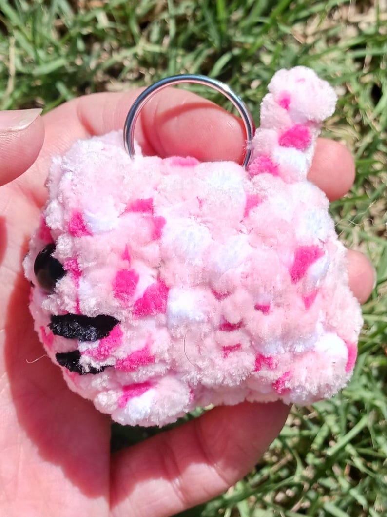 May include: A handmade, pink and white crochet keychain in the shape of a cat. The cat has black eyes and a black and white striped mouth. The keychain is held by a silver metal ring.