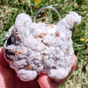 May include: A handmade, gray and brown crocheted cat keychain. The cat has black eyes and a silver keyring attached to its head. The plush toy is held in a hand against a background of green grass.