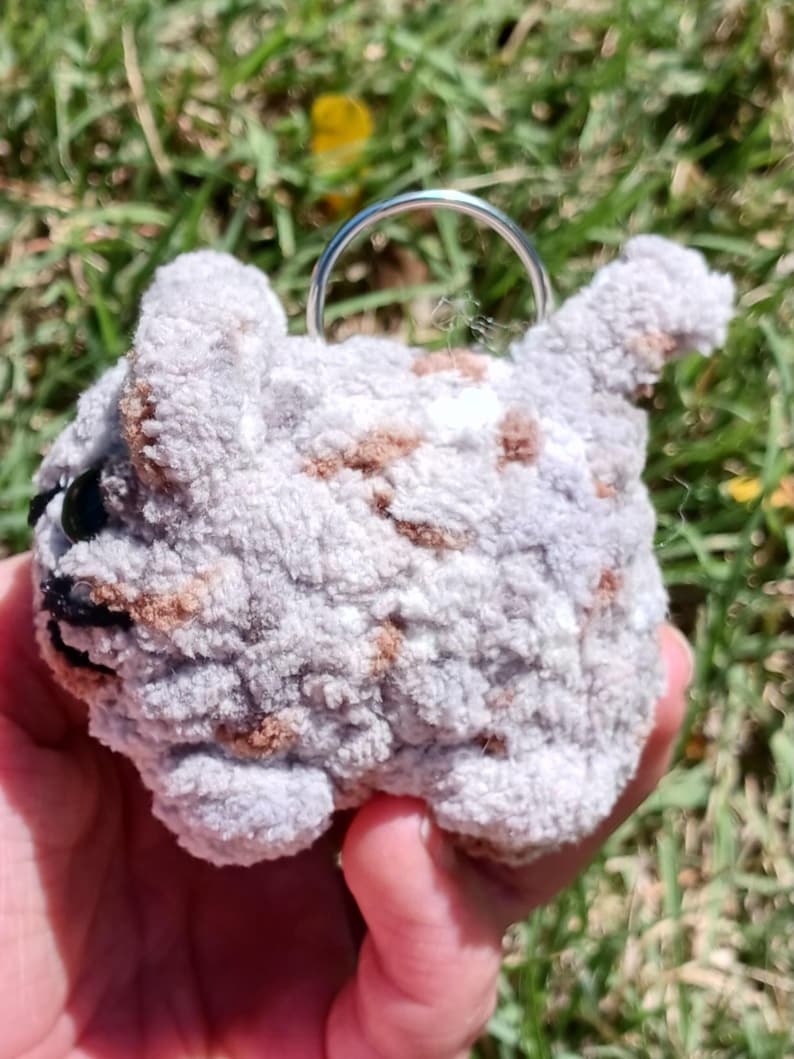 May include: A handmade, gray and brown crocheted cat keychain. The cat has black eyes and a silver keyring attached to its head. The plush toy is held in a hand against a background of green grass.