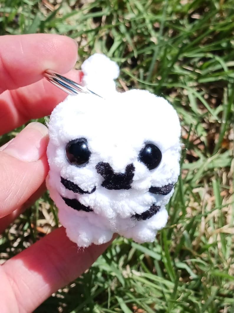 May include: A white, crocheted keychain featuring a small, round creature with black eyes and black markings. The plush toy has a fluffy texture and is held against a backdrop of green grass.