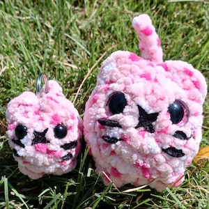 May include: Two plush cat toys in pink and white with black accents. The larger cat has a tail and the smaller one has a keychain. Both have black star-shaped eyes and black whiskers. The toys are sitting on green grass.