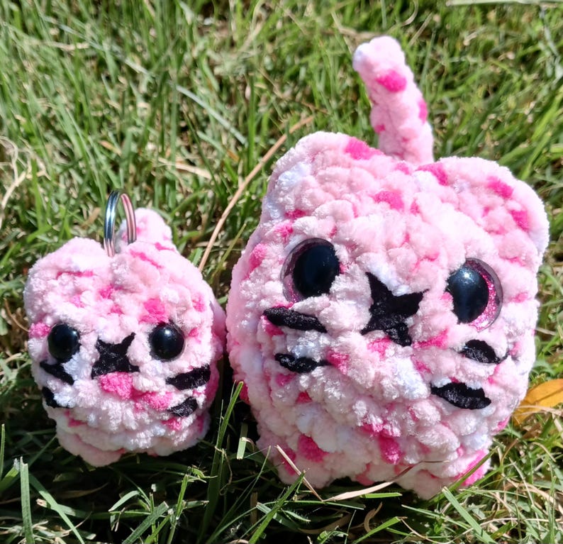 May include: Two plush cat toys in pink and white with black accents. The larger cat has a tail and the smaller one has a keychain. Both have black star-shaped eyes and black whiskers. The toys are sitting on green grass.