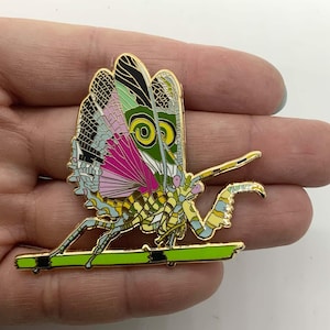May include: A gold-toned enamel pin featuring a colorful praying mantis with green, pink, and blue wings. The mantis is perched on a green branch.