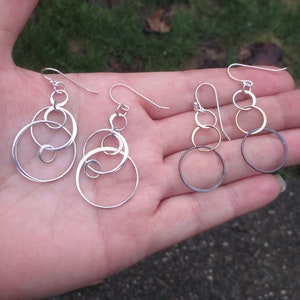 May include: A pair of silver and gold circle earrings with a dangling design. The earrings are made of multiple circles of different sizes, with the larger circles overlapping the smaller circles.
