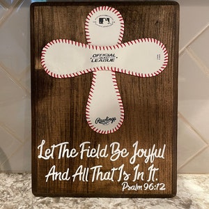 May include: A wooden sign with a white baseball stitched into the shape of a cross. The baseball has the words "Official League" and "Rawlings" printed on it. The sign also has the text "Let The Field Be Joyful And All That Is In It. Psalm 96:12" printed in white.