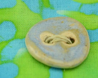 May include: Close-up of a ceramic button with a light blue and beige color scheme. The button has two holes with beige thread and is set against a vibrant green and blue patterned fabric.