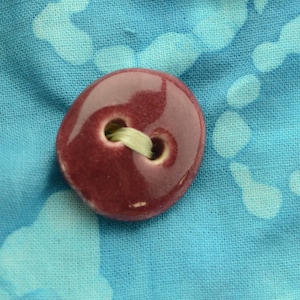 May include: A close-up of a burgundy ceramic button with two holes and a light green thread. The button is on a blue fabric with a white pattern.