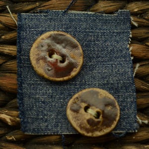 May include: Two brown ceramic buttons with a textured appearance are displayed on a small square of blue denim fabric. The buttons have a circular shape with a lighter-colored rim and a dark brown center. The denim fabric has frayed edges.