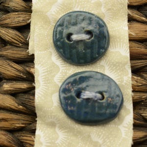 Könnte beinhalten: Zwei dunkelblaue Keramikknöpfe mit weißem Faden, befestigt an einem Stück hellgrünem Stoff mit weißem Blumenmuster. Die Knöpfe haben eine glänzende Oberfläche und sind vor einem gewebten Hintergrund angeordnet.