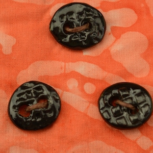 May include: Three glossy black buttons with a textured surface and two holes, each with a tan thread visible. The buttons are arranged on a coral-colored fabric with a white pattern.