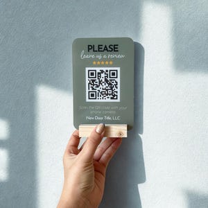 May include: A rectangular sign with rounded corners, featuring the text "PLEASE leave us a review" and a QR code. The sign is held by a small wooden block. The text "Scan the QR code with your phone camera" is also present.
