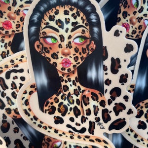 Könnte beinhalten: Ein Aufkleber mit einer Frau mit einem Leopardenmuster im Gesicht und am Körper. Ihre Augen sind grün und sie hat eine rosa Glitzerlippe. Der Aufkleber hat einen weißen Hintergrund und den Text "ART CGE" in der rechten unteren Ecke.
