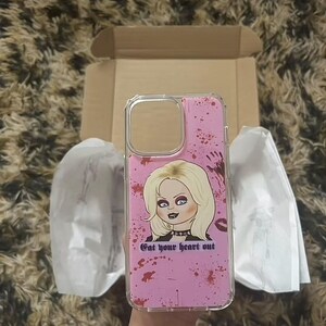 K&ouml;nnte beinhalten: Eine rosa Handyh&uuml;lle mit durchsichtiger R&uuml;ckseite und einem Grafikdruck einer Frau mit blonden Haaren und einem schwarzen Choker. Die Grafik ist von roter Spr&uuml;hfarbe umgeben und der Text "Get your heart out".