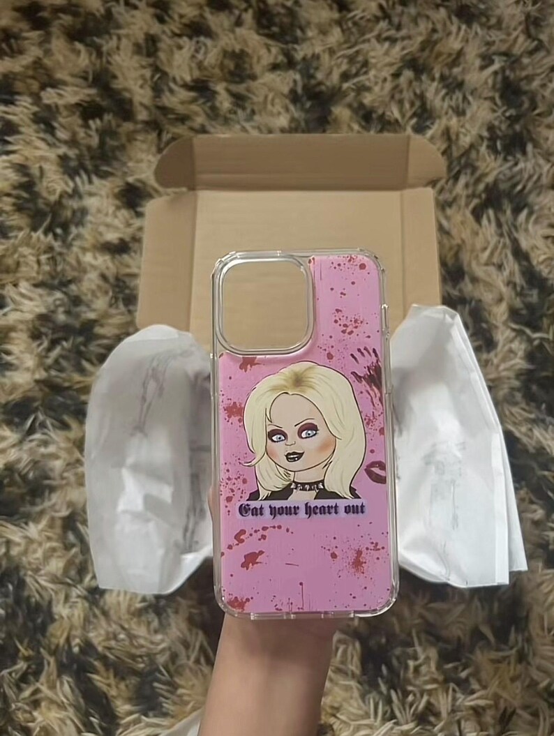 K&ouml;nnte beinhalten: Eine rosa Handyh&uuml;lle mit durchsichtiger R&uuml;ckseite und einem Grafikdruck einer Frau mit blonden Haaren und einem schwarzen Choker. Die Grafik ist von roter Spr&uuml;hfarbe umgeben und der Text "Get your heart out".