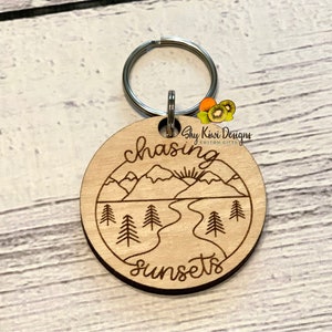 May include: Wooden keychain with a laser-engraved design of mountains, a river, and trees. The text "chasing sunsets" is engraved on the keychain.