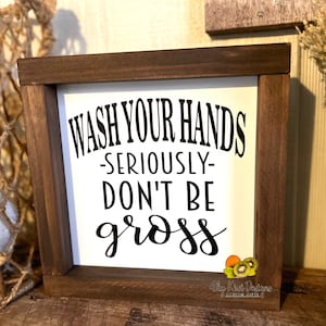 May include: A white wooden sign with a brown frame reads "Wash Your Hands -Seriously- Don't Be Gross". The sign is decorated with a small image of kiwi fruit and a sticker that reads "Shy Kiwi Designs".