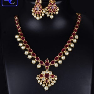 May include: A gold-toned necklace and earring set. The necklace features a series of oval, dark red stones interspersed with small, round, white pearls. The pendant has a cluster of red stones and pearls. The earrings mirror the necklace design. The text "Lc:109020 Cz Neckalce" is visible.