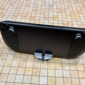 May include: A black handheld gaming device with two analog sticks, a D-pad, and buttons. The device is on a gray stand and has a black screen.