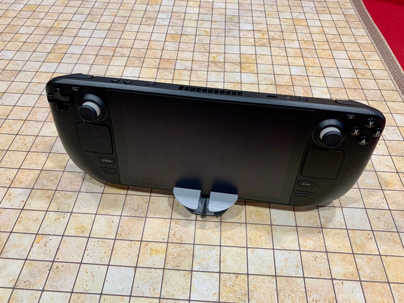 May include: A black handheld gaming device with two analog sticks, a D-pad, and buttons. The device is on a gray stand and has a black screen.