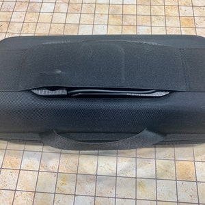 May include: Black hard-shell case with a zipper and a carrying handle. The case has a black strap across the top.
