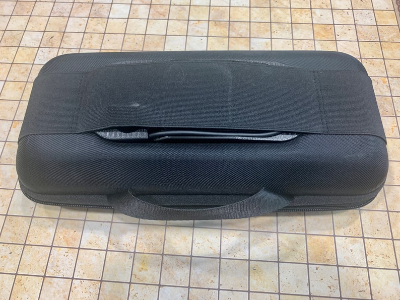 May include: Black hard-shell case with a zipper and a carrying handle. The case has a black strap across the top.