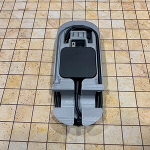 May include: A 3D printed gray holder for a black power adapter and cord. The holder has a slot for the adapter and a groove for the cord.