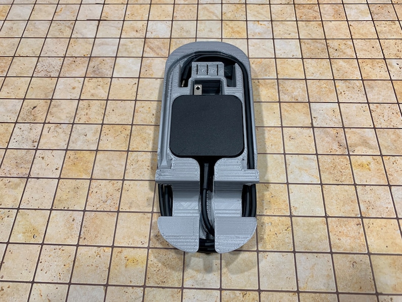 May include: A 3D printed gray holder for a black power adapter and cord. The holder has a slot for the adapter and a groove for the cord.
