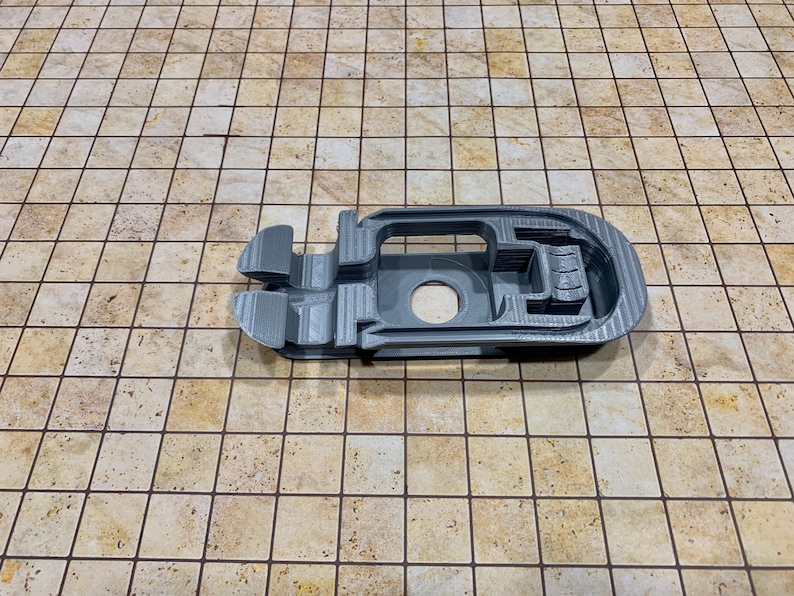 May include: A gray 3D printed plastic object with a complex design, featuring multiple openings and a circular hole. The object is lying on a beige tiled surface.